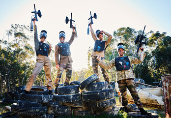 Team, paintball and portrait in celebration for winning, victory or achievement standing on tires together in nature. Group of people enjoying win, success or teamwork with guns in the air for sports