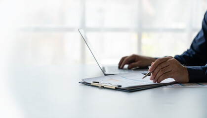 young business man Recording data on laptop, comparing details from graphs. Business income charts in the areas of real estate, tax, management, technology marketing in a private office.