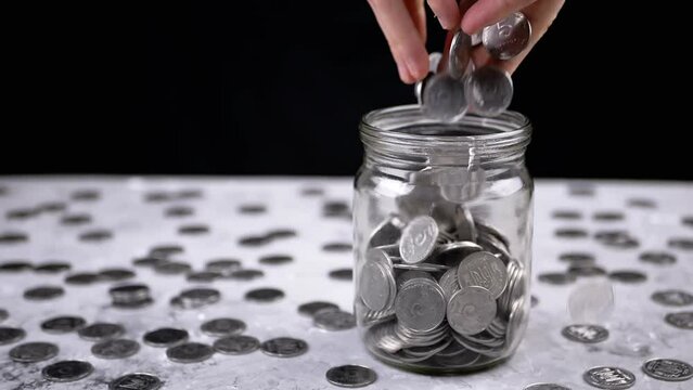 Hands Pouring a Handful of Accumulated Silver Coins into a Glass Jar. A Pile of scattered Ukrainian five kopecks is on table. Piggy bank. Black background. Financial crisis, budget savings. Poverty.