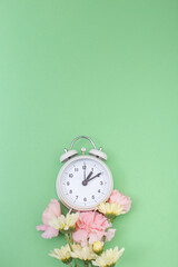 Reloj despertador color blanco ilustrando cambio de horario primavera, verano rcon base de flores naturales sobre fondo verde