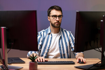 Serious handsome designer, editor working freelance project looking at computer monitor. Portrait of successful bearded computer programmer sitting at workplace, late night 