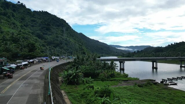 Rising Overhead Aerial Shot of tricycles and vehicles traveling on mountainside roadway leading to Bato Bridge amid vast river and lush jungles. Catanduanes, Philippines