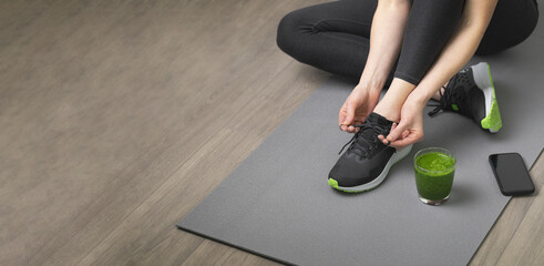 Women's hands tying sport shoes on a gray workout mat. With smoothie for detox in background....