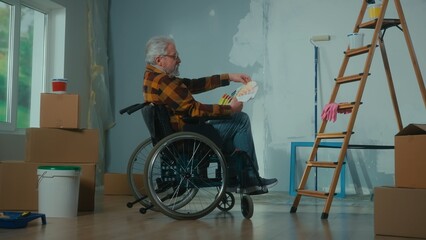 An elderly disabled man in a wheelchair looks through the palette with colors. A pensioner plans repairs and chooses a paint color. Room with window, ladder, cardboard boxes, wallpaper, paint.