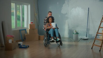 Fototapeta na wymiar A white man and African American woman in a wheelchair with a bucket of paint in her hands. Black woman with surprised face and shocked emotion. The concept of renovation in the apartment.