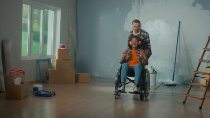 Fototapeta na wymiar A white man and African American woman in a wheelchair. Young couple in big headphones enjoy music, sing and laugh. Family on the background of the room in the process of renovation.