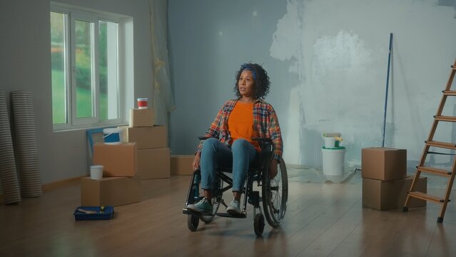 Portrait of young African American woman in a wheelchair. A disabled woman thinks and plans repairs in the room. Handicapped person on the background of cardboard boxes, cans of paint and a ladder.