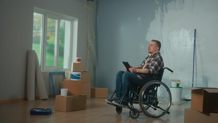 A young man in a wheelchair is planning repairs using a digital tablet. Disabled male browsing interior design ideas on the internet. Handicapped person.