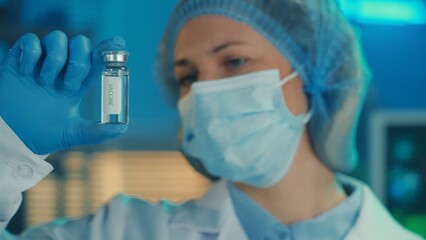 Portrait of a woman in a white gown, medical mask, bonnet and blue gloves holds a glass vial with a vaccine in her hand. A female doctor examines the vial, blue light. Close up.