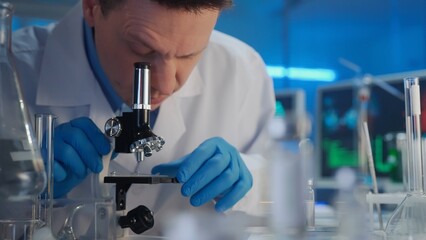 A male researcher in a white gown and blue gloves is examining a sample in a microscope. Laboratory...