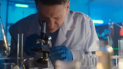 A male researcher in a white gown and blue gloves is examining a sample in a microscope. Laboratory or hospital with many test tubes and glass flasks. In the background is a computer monitor.