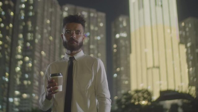 Medium shot of young handsome businessman in formal outfit and glasses holding to go coffee cup and posing for camera outdoors in night city