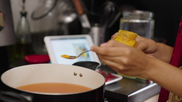 South asian woman putting spoonful of turmeric in pot of golden curry.