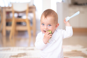 Baby with blue eyes and white bodysuit sit and eat cucumber, hold spoon. Child eating first food. Problems for feeding up children for parents. Healthy and unhealthy ready breakfasts.