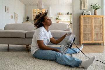 Tired overheated black woman cooling herself with electric fan at home, suffering from heat while sitting on floor in living room without conditioner, keeping cool indoors during summer heatwave - obrazy, fototapety, plakaty