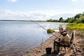 Obraz na płótnie Canvas Side view of bearded fisherman in raincoat sitting on river bank on travel chair with fishing rod waiting for catch on summer sunny day sandbank. Concept of lifestyle, leisure activity on nature