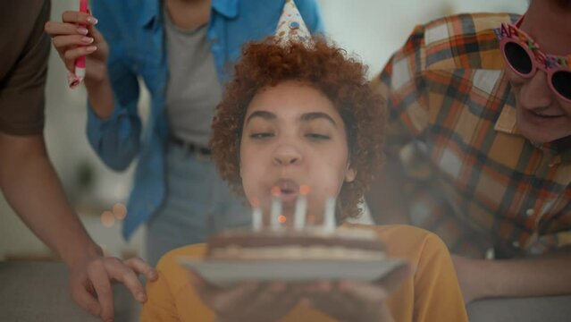African american girl celebrates birthday with friends blows candles on cake makes a wish. They smile and clap hands. Wearing festive caps and blows party horns. Celebration party for birthday girl.
