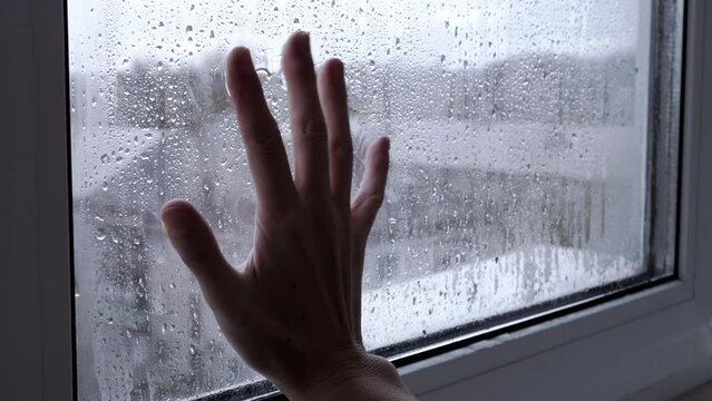 Woman Touches a Wet Misted Window with Hand. Fingers slide on wet glass. Drops of the condensate flow down. Isolation is at home. Gloomy rainy day. Hope, separation, despair, longing, loneliness.