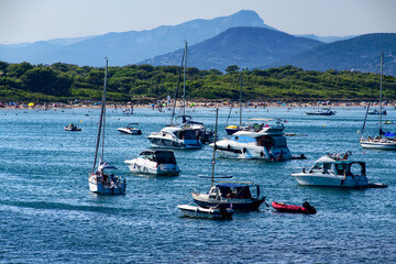 Fototapeta na wymiar A majestic sailboats and pleasure boats lays in front of the beach of Plage La Bandine, surrounded by sea and mountain views.