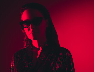 Young beautiful confident brunette female in trendy evening shiny green reflecting dress. Sexy carefree woman isolated on red neon light in studio. Fashionable model in sunglasses. high fashion
