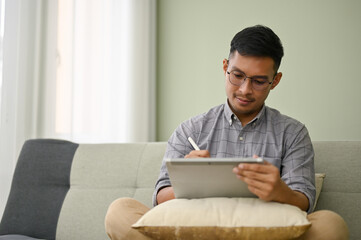 Focused millennial Asian man using his digital tablet on sofa in his living room.