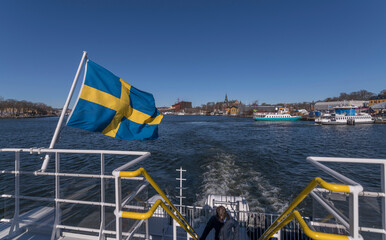 Waving Swedish flag on a harbor commuting ferry, blurred background with ferries, museums, a sunny spring day in Stockholm