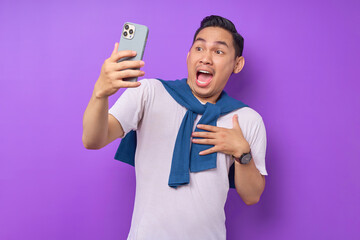 Surprised young Asian man using smartphone, reading special offer isolated over purple background