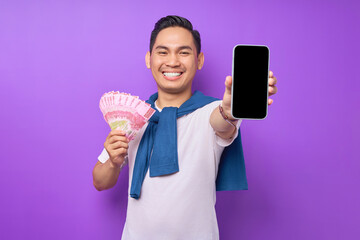 Cheerful young Asian man holding mobile phone with blank screen and money banknotes isolated over...