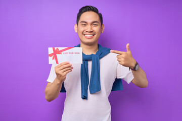 Smiling young Asian man in casual clothes pointing a finger at gift voucher certificate and looking at the camera isolated on purple studio background