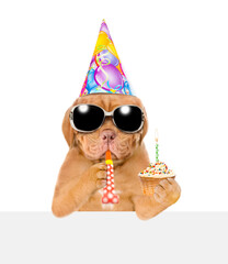 Mastiff puppy wearing sunglasses and party cap blowing in party horn holds birthday cupcake with burning candle above empty white banner. isolated on white background