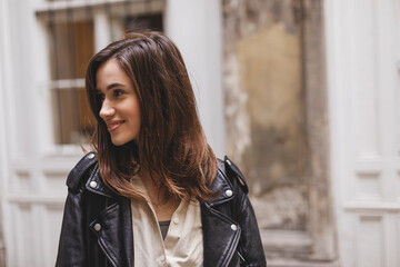 Trendy girl with brunette hairstyle posing outside. Woman in leather jacket and shirt look at size walking on street outdoors. Tourist happy woman posing in the city. Optimistic lady walks outside.