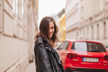 Naklejka na ściany i meble Trendy girl with brunette hairstyle posing outside. Woman in leather jacket and shirt walking on the street outdoors. Tourist happy woman posing in the city. Optimistic lady turn around.