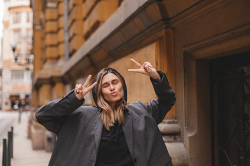Cool blonde woman in black hoodie and grey jacket shows peace sign, blows kiss and posing outside. Happy woman walk and posing outdoor.
