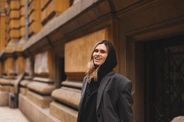 Fototapeta premium Trendy girl with blonde hairstyle posing outside. Woman in grey jacket and hoodie walking on the street outdoors. Tourist happy woman posing in the city. Optimistic lady walks outside.
