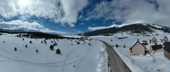 Aerial, drone shot over leafless trees, above a snowy mountain and beautiful snowy mountain peaks. Winter wilderness, on a sunny day. 