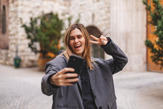 Happy young woman photographing herself using her mobile phone. Caucasian female taking selfie with her smart phone at city. Positive lady making photo outside in grey jacket and show peace sign.