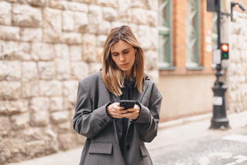 Photo of young woman reading messages on a smartphone while going on the street. Blonde caucasian...