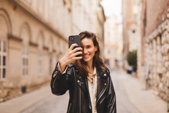 Happy woman photographing herself using her mobile phone. Caucasian female talking selfie with her smart phone at city. Positive lady making photo outside in leather jacket. Focus on phone.