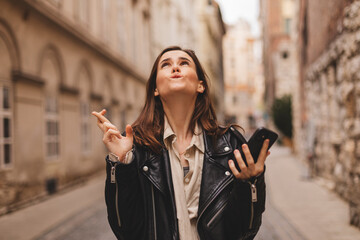 Portrait of wishful young woman in casual clothes with brunette hair, crossing her fingers, feeling nervous before important event, walking on the street. Body language.