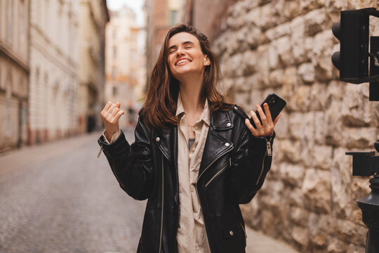 Delighted brunette girl making winner gesture and using mobile phone walking on the street. Portrait of surprised hipster girl surfing internet on mobile phone outdoors.