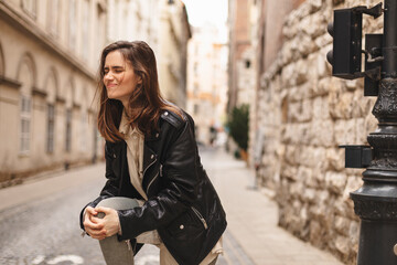 Fototapeta premium Slim brunette woman hurting knee joint while walk on the street. Girl hand in ache knee in street.