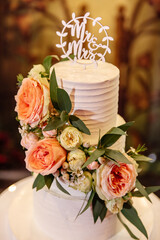 A handmade wedding cake decorated with large pink flowers and green leaves sits on a white cake table. The top of the cake is decorated with the inscription Mr. and Mrs. in white.