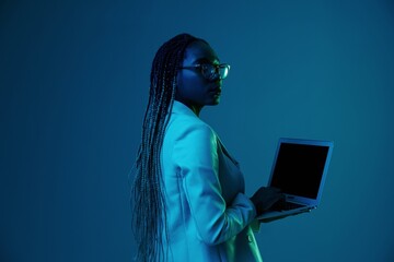 Naklejka na ściany i meble African African woman with laptop in glasses on a blue background in neon light, color mixed light. Data storage cyber security, hacker, big data.