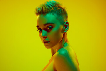 Beautiful young girl with short blonde hair posing with bare shoulders against yellow studio background in green neon light. Cyberpunk style. Concept of technology, modern fashion, digital art