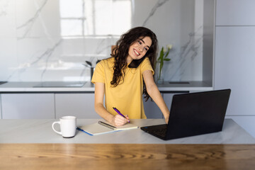 Portrait of a young woman talking the phone and working on laptop in kitchen