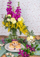 Tischdekoration im Frühling mit frischen Blumen und Muffins, Blumenstrauß und Gepäck auf einem Tisch