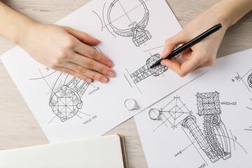 Jeweler drawing sketch of elegant ring on paper at wooden table, top view