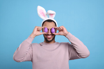 Happy African American man in bunny ears headband covering eyes with Easter eggs on light blue background