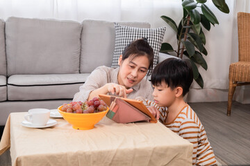 Mother teaching handsome kid to review lessons by tablet during the holidays, woman and son sit on warm floor and learn lesson together in living room. domestic life and family leisure time concept.