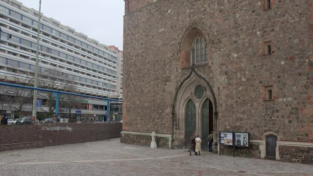 People enter the church building. A small door in a high wall. City street, pedestrian traffic. Tall long building in the background. 